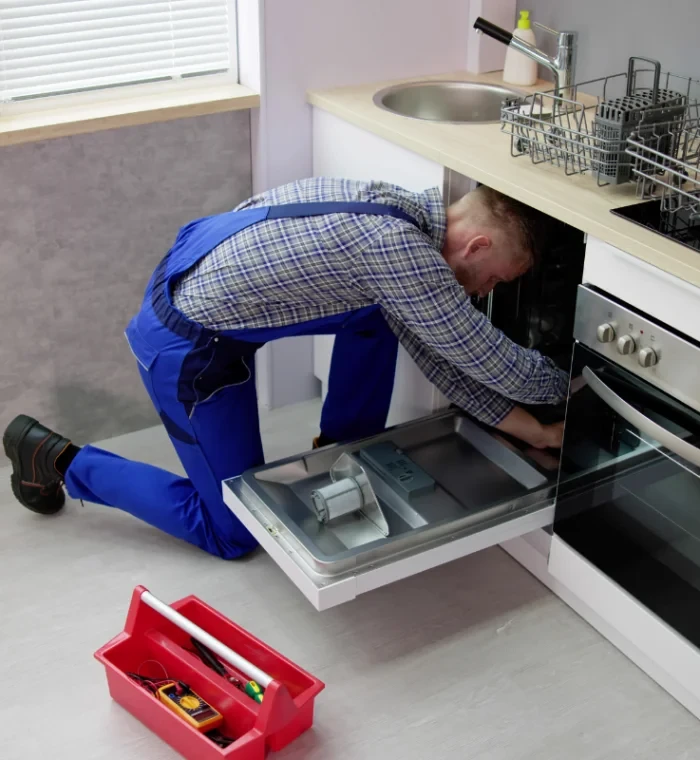 Technician servicing a dishwasher beside an oven with a red toolbox nearby